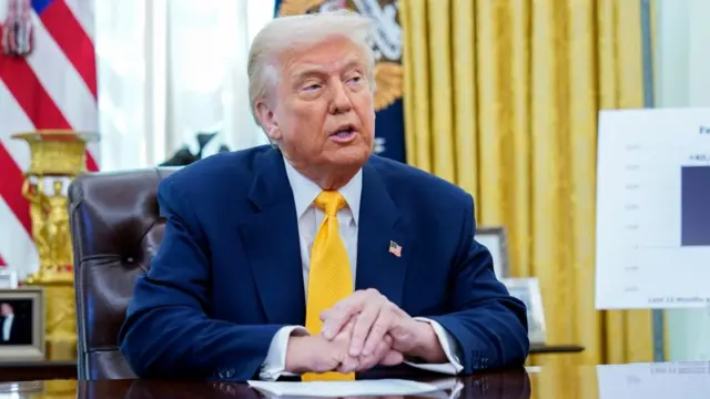 Donald Trump speaks behind a desk in the Oval Office, wearing a blue suit and yellow tie.