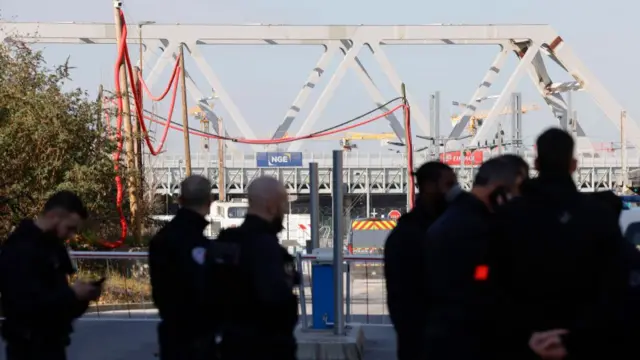 Police officers stand in the foreground with infrastructure for moving freight is visible in the background