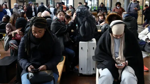 A large crowd of passengers, with luggage, sit on benches looking at their phone.