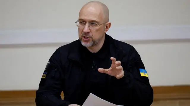 Ukraine's Prime Minister Denys Shmyhal speaks at a press conference, wearing a black top with the Ukrainian flag on it.