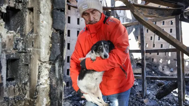 A man in a red jacket carries a small black and white dog from a house in Odesa