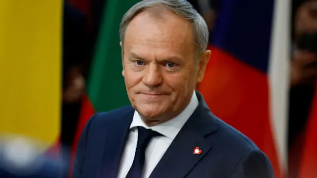 Poland's prime minister Donald Tusk, wearing a dark suit and tie, with blurred flags in background, shot from chest up.