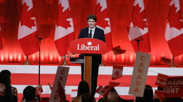 Justin Trudeau delivers speech at lectern with sign reading Liberal at the front. A row of Canadian flags behind him as a crowd waves placards reading Canada