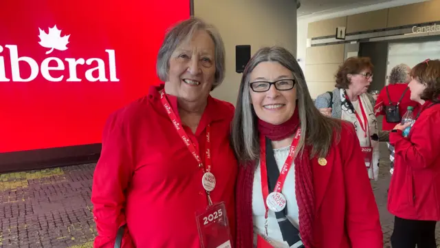 Janet Conn and Sophie Kiwala stand side by side. Both wear bright red, which is the colour of the Liberal Party of Canada, and they are smiling. Behind them is a sign with the Liberal logo.