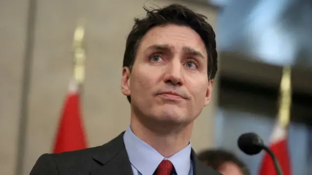 Close up of Justin Trudeau as he delivers speech in dark grey suit, light blue shirt and red polka dotted tie. The top of two golden flag poles are visible in the background