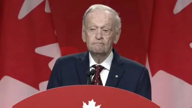 Jean Chretien delivers speech at lectern in a dark blue suit, white shirt and burgundy tie, a row of Canadian flags behind him