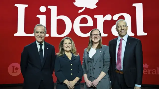 Mark Carney, Chrystia Freeland, Karina Gould and Frank Baylis stand in a row smiling in front of a sign reading "Liberal" during a leadership debate in February