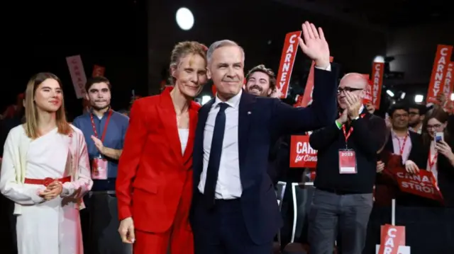 Mark Carney (R) hugs his wife as he waves at the crowd following his election. He's standing in front of a crowd, his daughter Cleo in a white shirt and trousers looks at them from the left of the frame