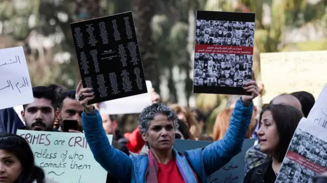 A woman holds up two signs, one with the names of those reported to have been killed and another with their pictures