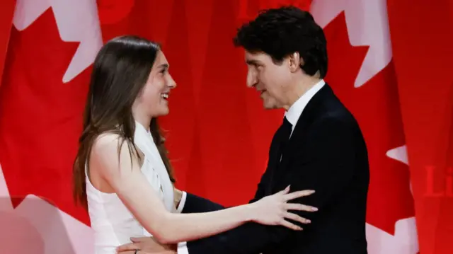 Ella Grace Trudeau (L) in a white jumpsuit greets Justin Trudeau (R) to the stage, Canadian flags in the background