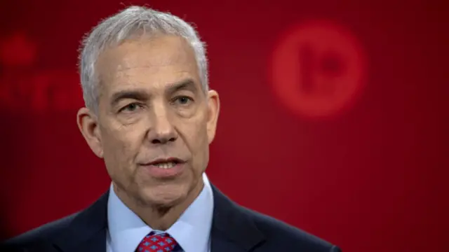 Frank Baylis (middle-aged man with short-cropped grey and white hair) speaks on podium wearing a dark blue suit, a red wall in the background