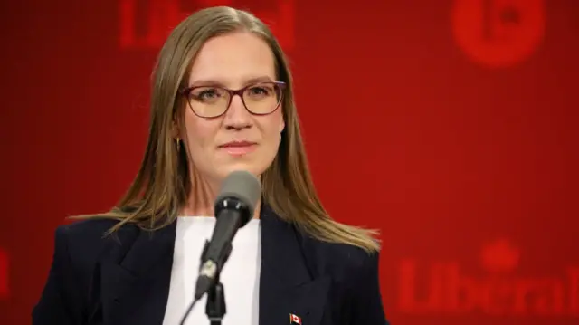 Karina Gould close up as she stands in front of a mic in a black blazer and white shirt. She has long light brown hair and is wearing glasses.