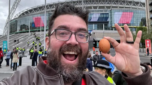 Kevin Chapman a man who is standing in front of Wembley stadium. He has dark hair and facial hair and is wearing glasses. He is looking directly at the camera and smiling with his mouth wide open. He is wearing a brown jacket and is holding an egg up to the camera.