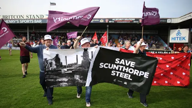 Steelworkers, members of UNITE Union and supporters hold banners and flags as they walk on the pitch at Attis Arena home of National League North team Scunthorpe United