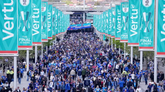 Thousands of fans in blue and white shirts are walking up Wembley way towards the Wembley stadium. There are dozens of teal flagpoles on either side of the path, with the word "Vertu" on them