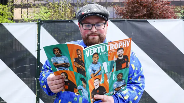Fan at Wembley wearing a flat cap who is wearing a blue jacket and holding a Vertu Trophy magazine
