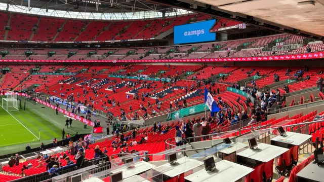 Fans are getting into their seats inside Wembley. The seats are red and there is a blue and white flag being held up in one of the stands
