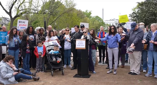 A group of people standing around with signs saying bring Kilmar home. A woman stands at a lectern with a microphone speaking