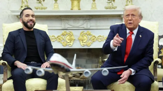 Bukele and Trump sitting on chairs in a fancy room with a big fireplace behind them. Trump points in front of him and speaks as Bukele smiles