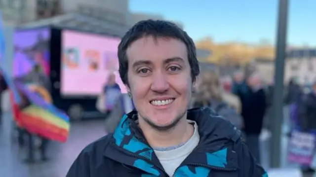 Vic Valentine smiles at the camera. Vic has short dark hair and wears a dark jacket with blue sections. They stand in front of a demonstration outside of the Scottish parliament.