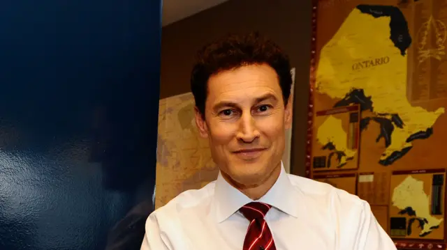 Steve Paikin, wearing a white shirt and a burgundy and white tie, leans on a navy blue background and smiles at the camera. A yellow coloured map of Ontario is behind him, right of screen..