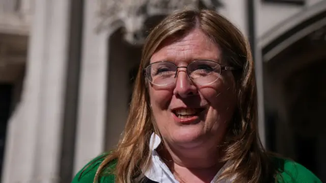 Susan Smith wears a white shirt and green blazer and smiles outside the Supreme Court building