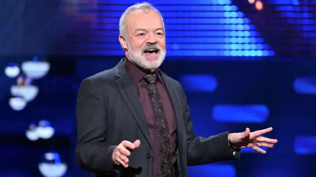 Graham Norton during the filming for the Graham Norton Show at BBC Studioworks. He's in a black suit, aubergine purple shirt and patterned tie