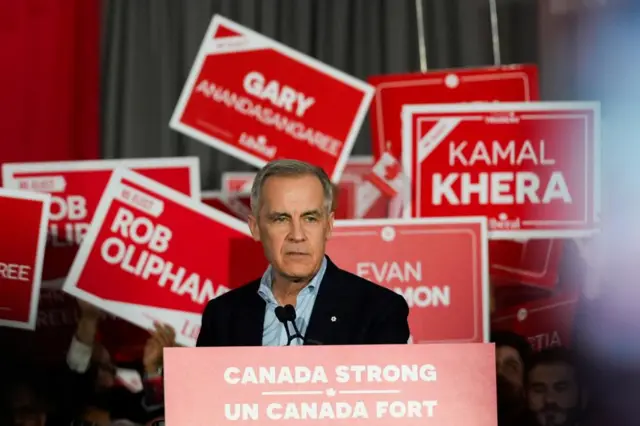Mark Carney stands behind a podium with a red banner in front. The banner has the words: Canada Strong and Un Canada Fort. Behind Carney, supporters hold up signs for other Liberal Party candidates including Kamal Khera, Rob Oliphant and Gary Anandasangaree.