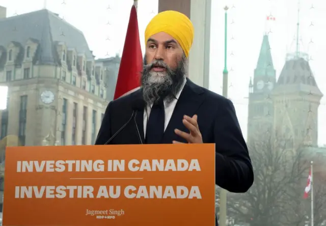 Jagmeet Singh speaks at a podium with an orange banner in front. The banner has the words: Investing in Canada on top and Investir au Canada on the bottom. He is wearing a black suit over white shirt and a tie and a yellow turban or dastar.