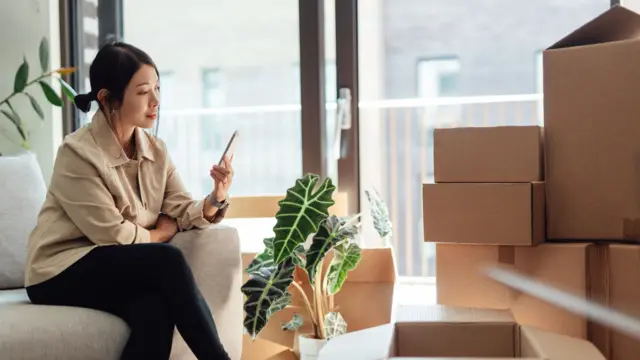 Asian woman with brown hair pulled back in a low bun sits down on a light grey couch checking her phone. In front of her is a stack of boxes including one containing a green plant