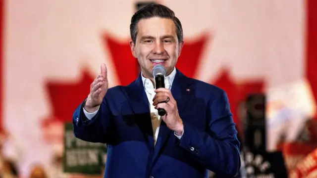 Pierre Poilievre, wearing a blue suit over a white shirt, speaks on stage. He is holding a microphone in his left hand and has his right hand outstretched forward. He is also wearing a Canada flag pin on his blue suit.