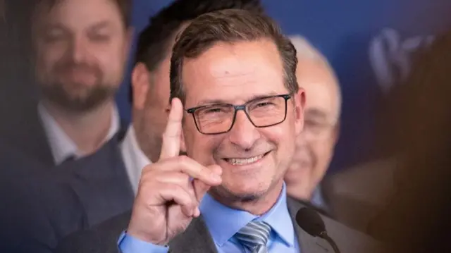 Yves-François Blanchet gestures with his right index. He is wearing a grey suit over a blue shirt and a silver-stripped tie.