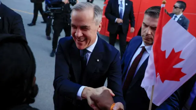 Carney smiling and shaking the hand of a person you cannot see. There's a red bus behind him and a small Canada flag on a stick being waved in his face