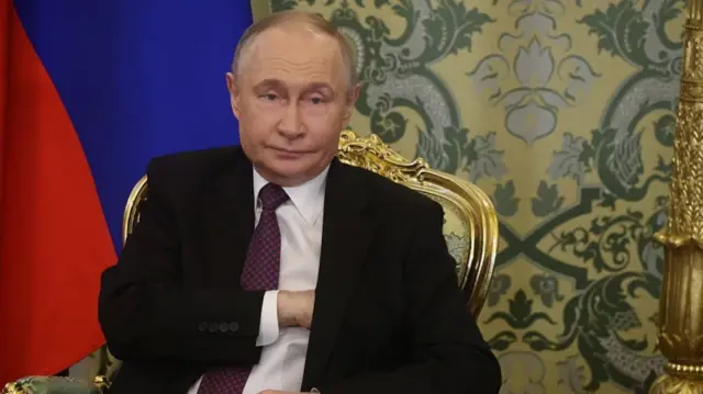 Vladimir Putin in a black suit, white shirt and deep burgundy tie sits on a wooden chair with golden details. A partial Russian flag is behind him