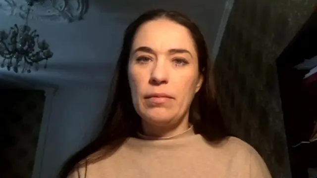 A woman looks ahead, wearing a beige jumper, in a house