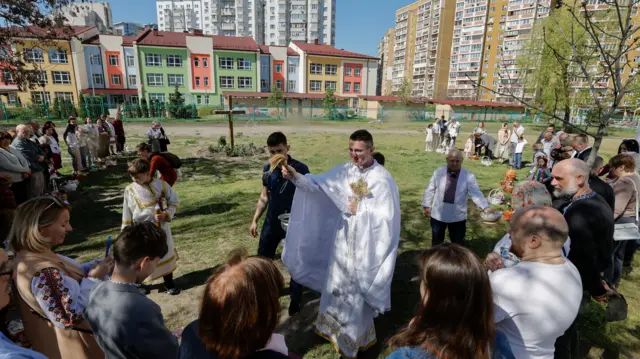 A priests holds a cross and blesses people standing round him with holy water