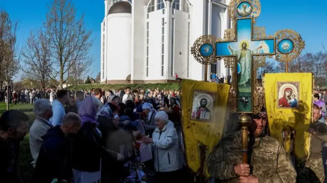 A man dressed in an army uniform holds a decorated cross. Crowds of people can be seen behind him. Part of a white church can be seen in the background