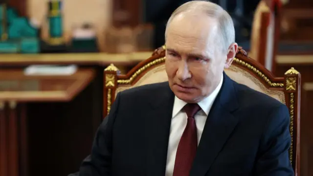 Russian President Vladimir Putin wears a suit while sitting on a chair and looks ahead