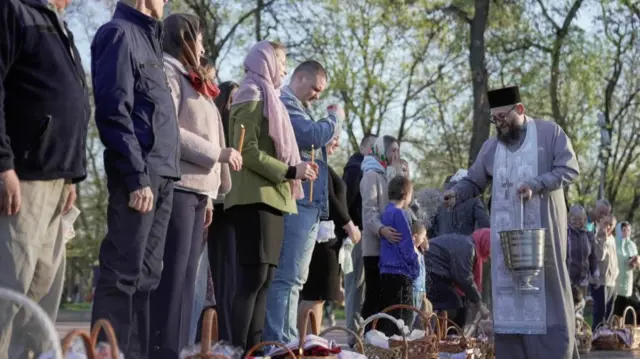 An orthodox priest carries a silver bucket as he sprays holy water onto baskets as people look on and hold candles