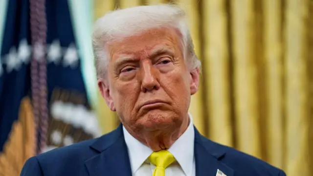 Donald Trump wears a suit, shirt and tie, and looks to the side in the Oval Office.