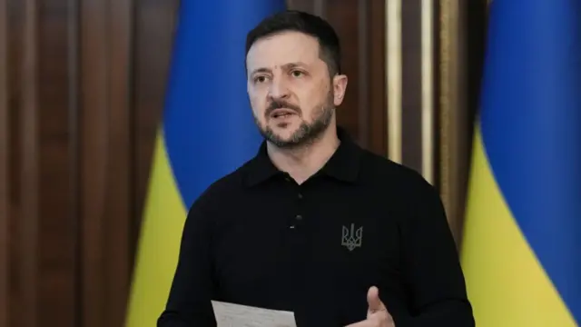 Volodymyr Zelensky stands at the centre of the frame in a black long-sleeved polo shirt while holding a piece of paper. Behind him are two Ukrainian flags