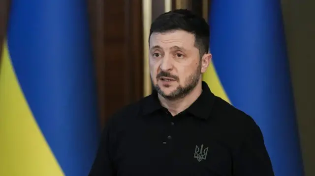 Volodymyr Zelensky in black long-sleeved polo shirt mid-speech as he stands with two Ukrainian flags at his back