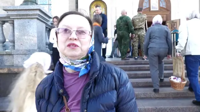 Olga wears a blue and white silk scarf around her head and purple top. She is standing outside church on Easter morning