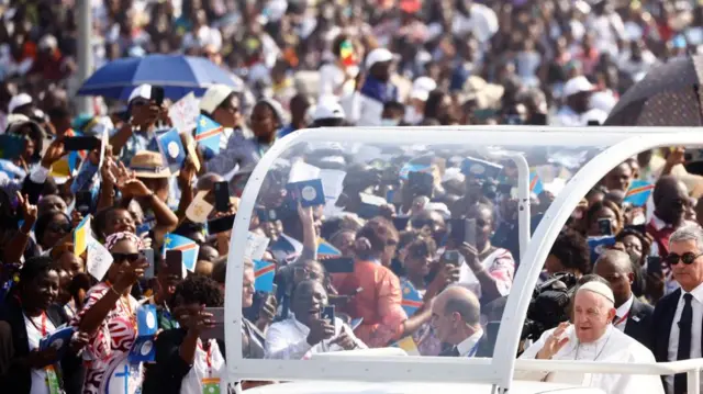 Pope Francis is being driven in a vehicle as large groups behind take pictures of him