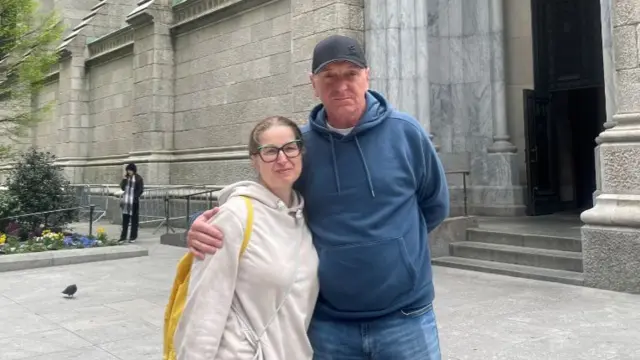 Andrea Sucha wearing a cream hoodie and a yellow backpack is pictured with her husband standing next to her who is wearing a blue hoodie and denims