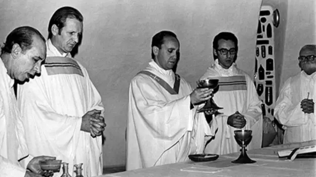 A black and white image of Bergoglio raising a cup over an altar surrounded by four other men