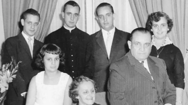 A black and white family photo of Francisco Bergoglio with his two brothers, two sisters and parents. He stands at the back of the frame with his brothers and older sister, his parents sitting on the couch with his younger sister
