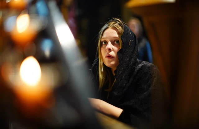 A woman wearing a black shoal looks above the camera as candles twinkle in the frame