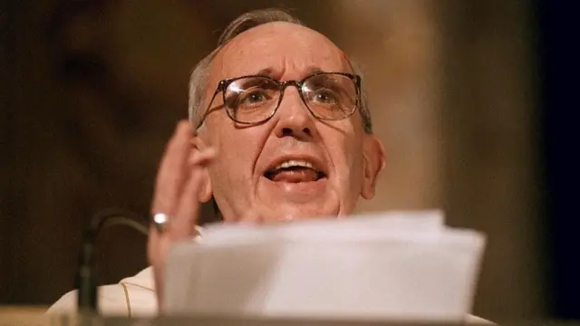 Close up of Bergolgio gesturing with his right hand while holding a stack of paper with his left as he gives a sermon