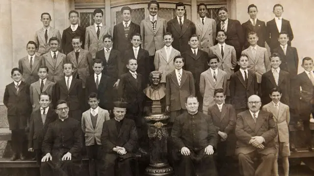 A black and white school photograph. All the pupils are male and wear suits and ties, and stand in three lines, with four teachers seated in the front row